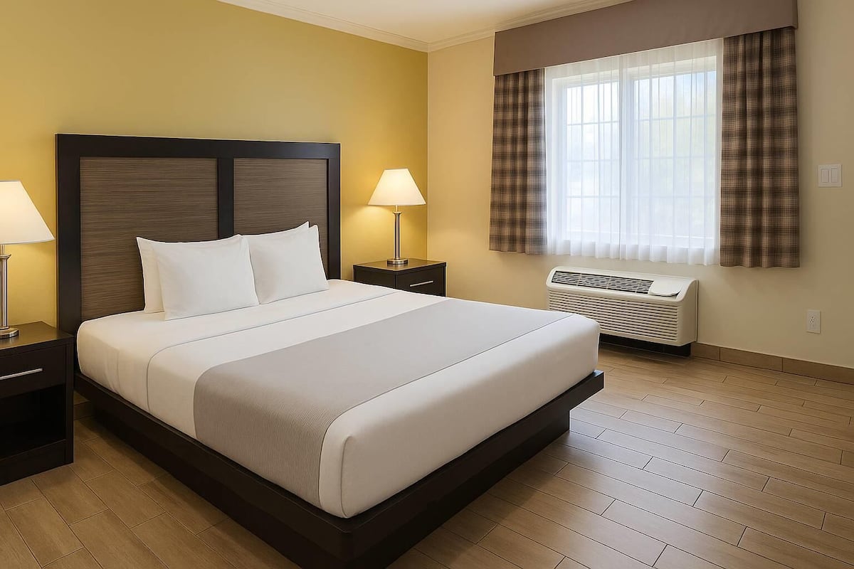 Una habitación de hotel con una cama grande, dos mesitas de noche con lámparas, una ventana con cortinas y un aire acondicionado instalado en la pared.