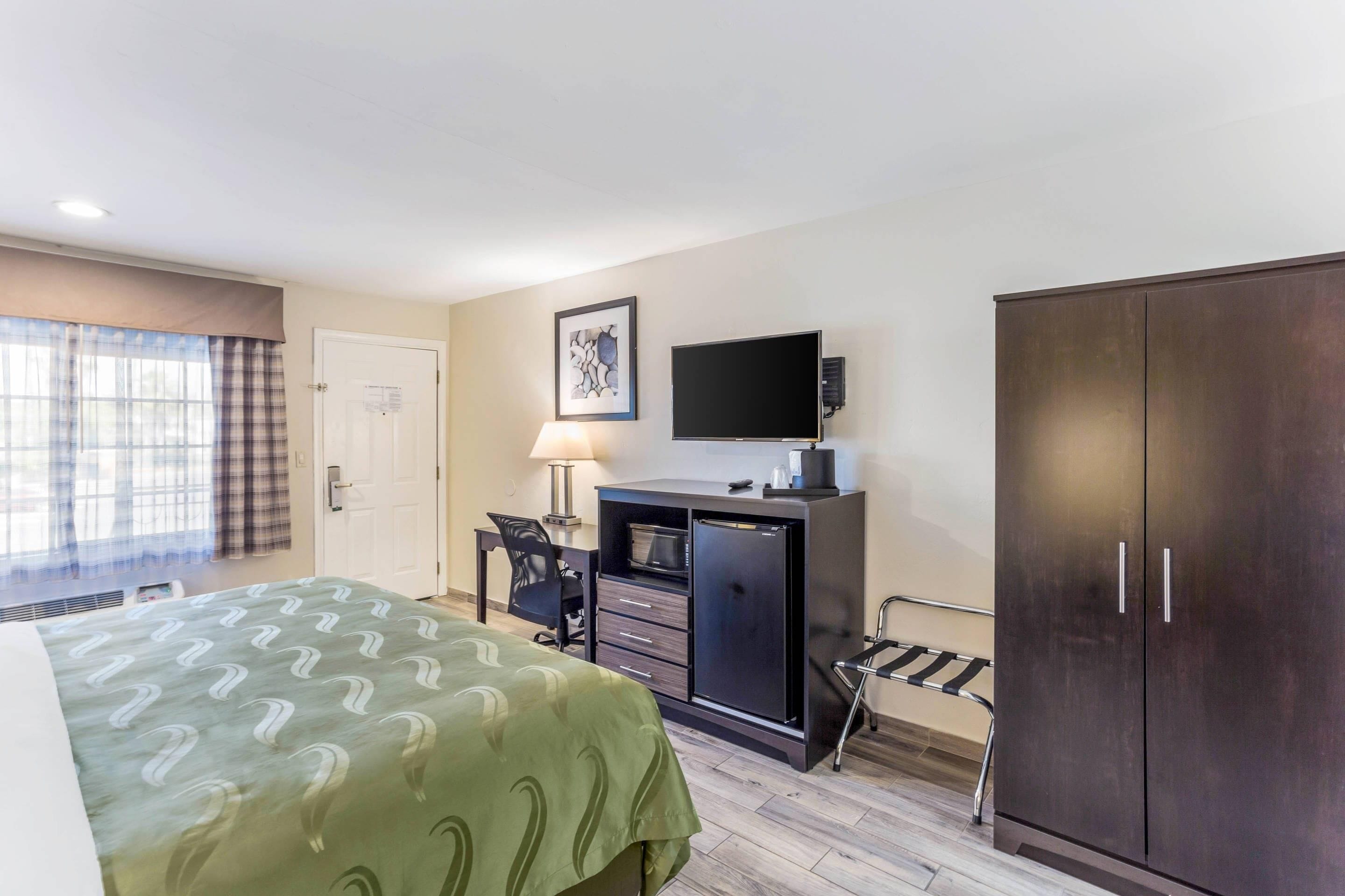 A hotel room with a bed, a desk with a lamp, a television, a wardrobe, and a window with curtains.