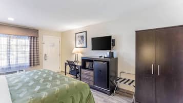 Habitación de hotel con cama, escritorio con lámpara, televisor, armario y ventana con cortinas.