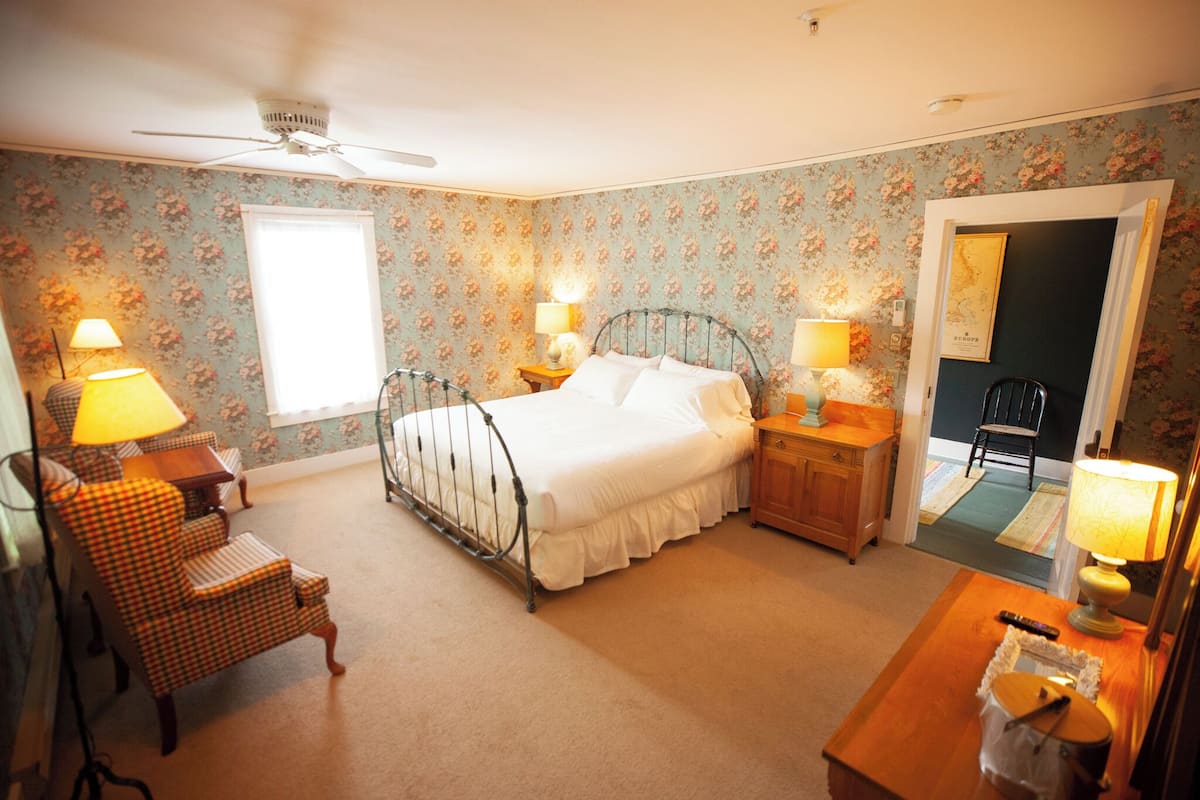 A bedroom with a bed, two bedside tables, a chair, a desk, a ceiling fan, and a door leading to another room.