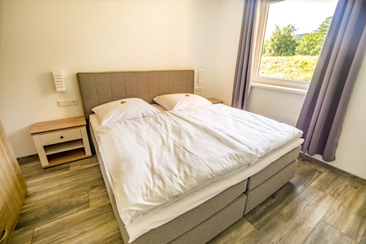 Una cama bien hecha con cabecera, una mesita de noche y una ventana con cortinas.