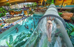 A water slide with a person at the top, a pool area with people, and a ship-themed structure. - AquaMagis (Plettenberg)