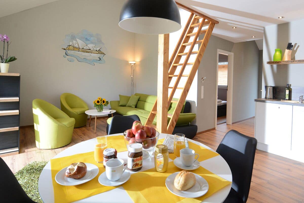Un comedor con una mesa lista para el desayuno, una escalera y una cocina al fondo.