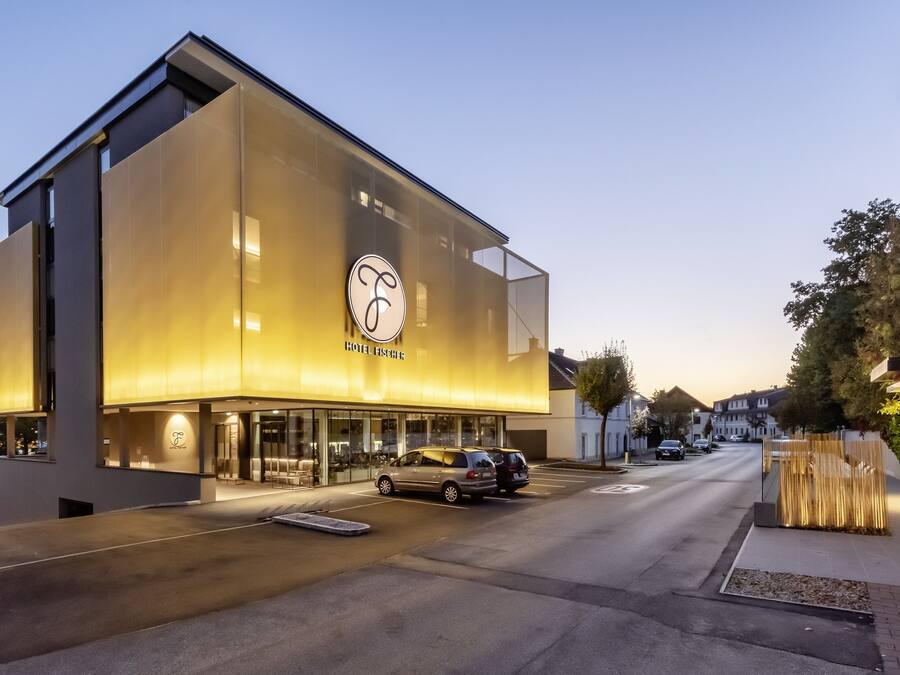 Un hotel moderno con un amplio frente de vidrio, un logotipo visible y un estacionamiento con automóviles.