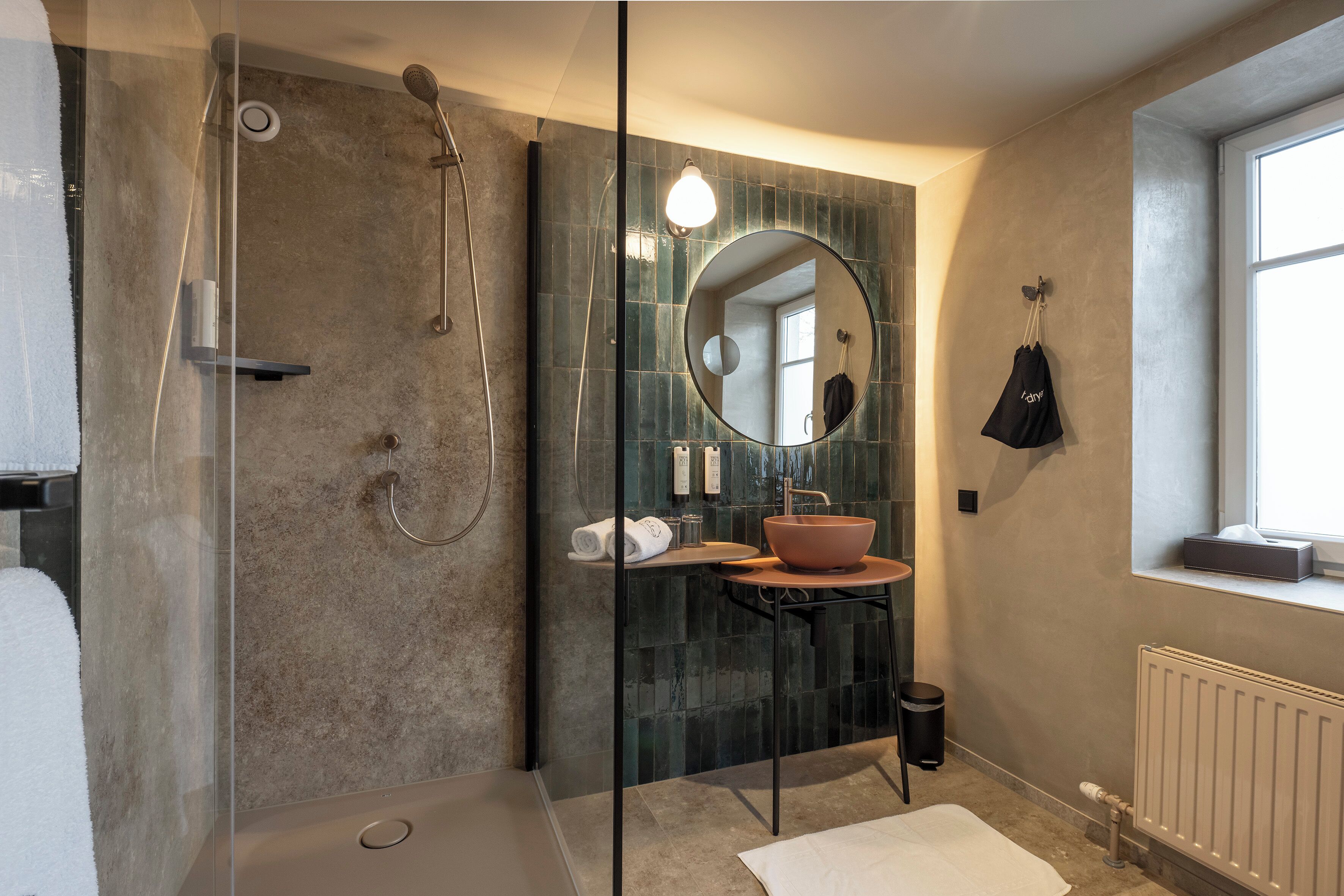 Un baño moderno con ducha envuelta en vidrio, espejo redondo, lavabo con recipiente y perchero para toallas.