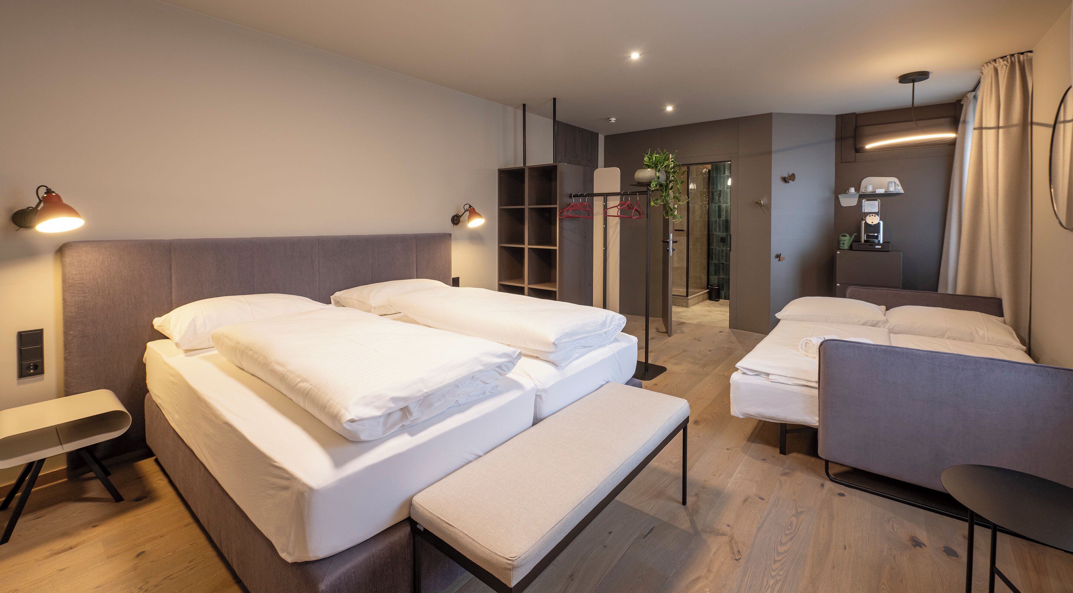 Ein Hotelzimmer mit Bett, Sitzbank, Stuhl, Nachttisch, Spiegel, Duschbereich und Kleiderschrank.