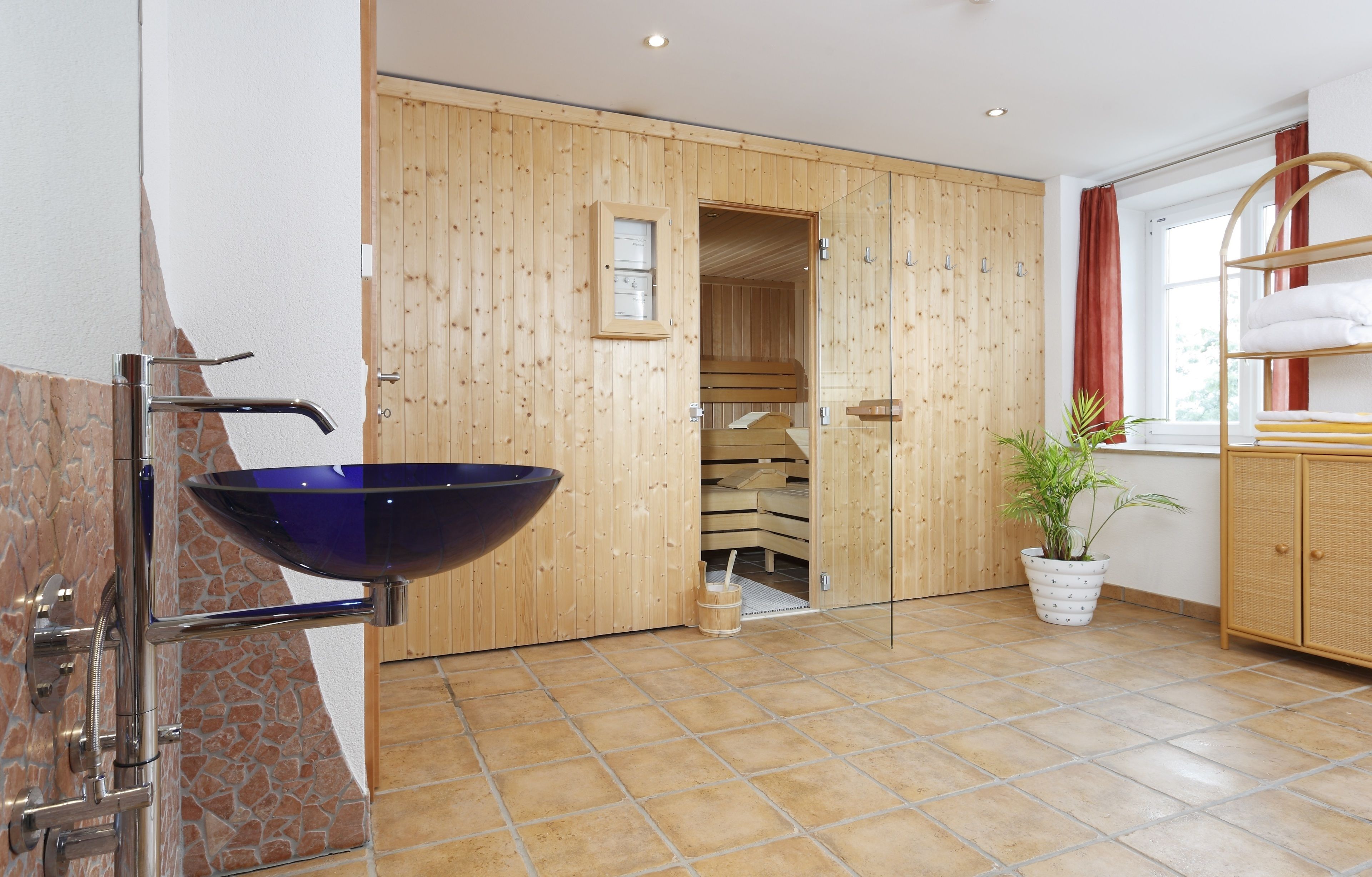 A modern bathroom with a large blue basin, wooden panel walls, a wooden sauna, and a potted plant.
