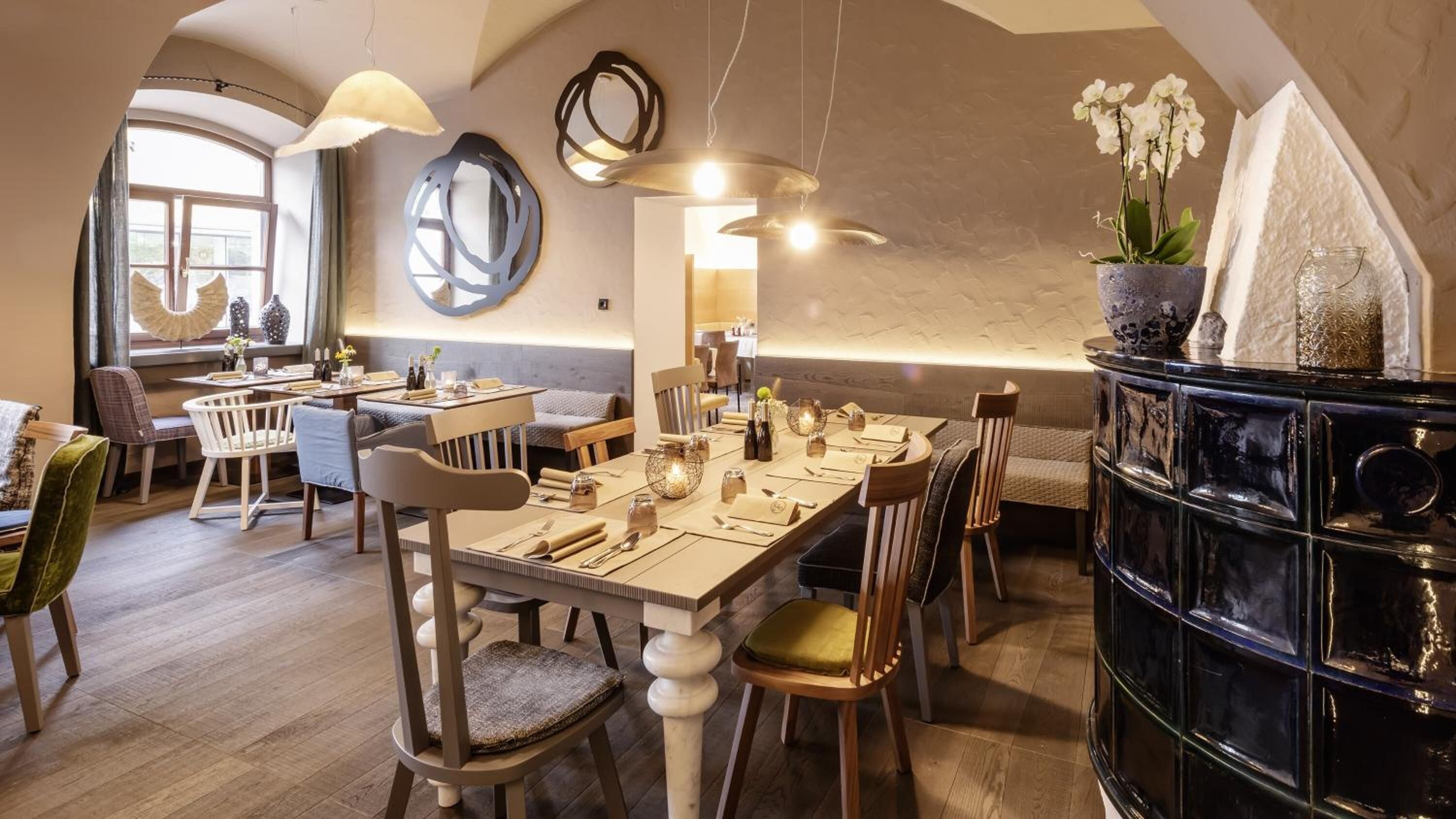 A dining area with a long table set for multiple guests, surrounded by chairs. There are hanging pendant lights above the table, and a decorative wall piece is visible.