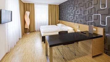 A hotel room with a wooden floor, a bed with white bedding, a desk with a black surface, a TV mounted on the wall, and a patterned wallpaper.