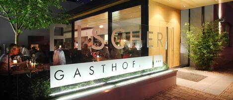 A restaurant with a sign reading "GASTHOF." It features an outdoor seating area with tables and chairs, a tree, and a well-lit entrance.