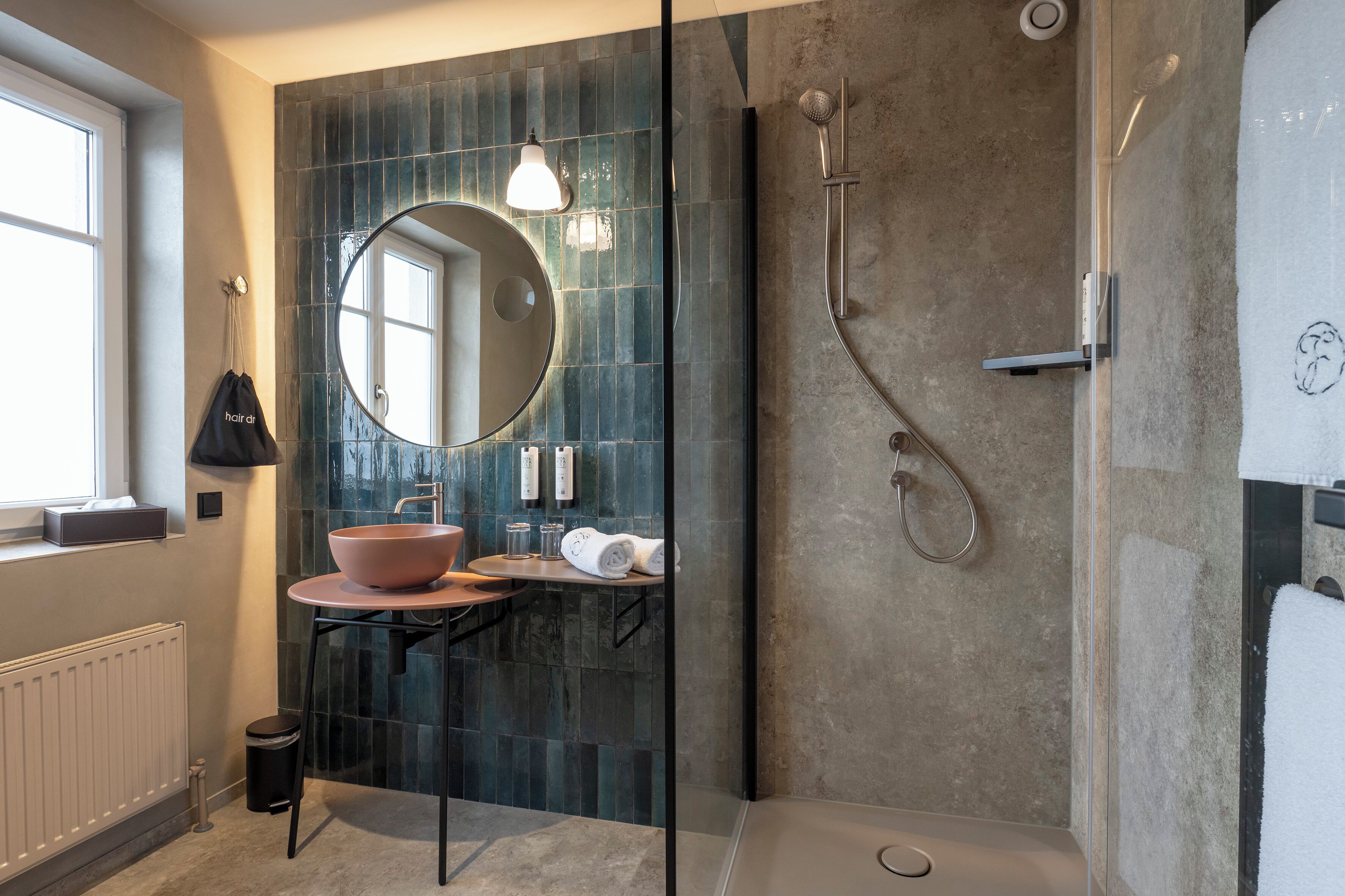 A modern bathroom with a round mirror, a pink sink, a shower, and a towel rack.