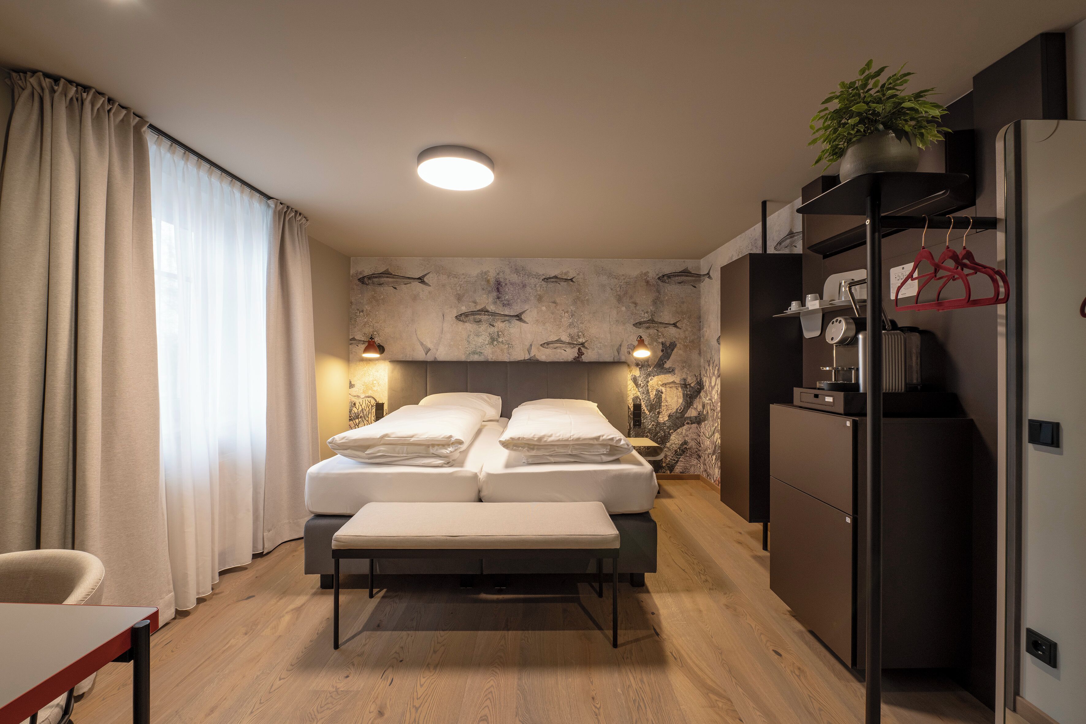 Una habitación de hotel con cama, mesitas de noche, un banco, un escritorio, un refrigerador y una pared con motivos de peces.