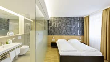 Quarto de hotel moderno com box de vidro, piso de madeira, cama com roupa de cama branca, um espelho grande e parede com padrão.