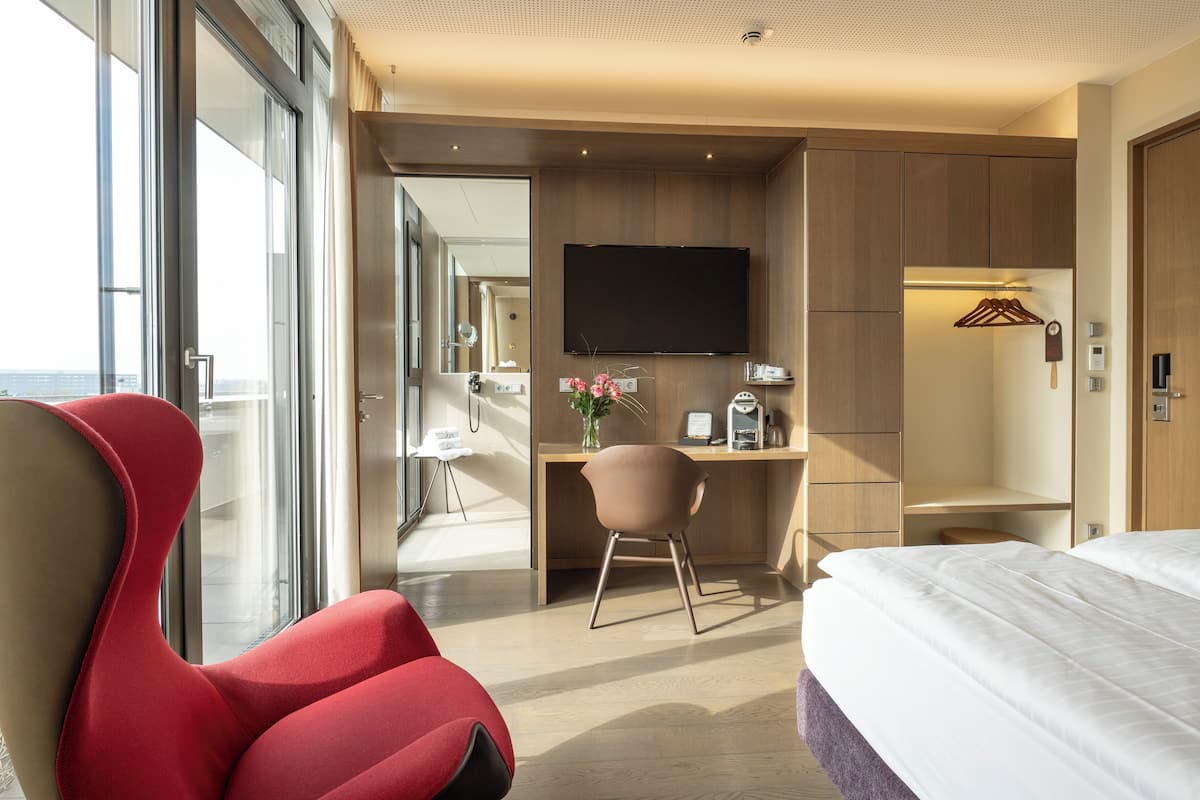 A modern hotel room with a large bed, a red armchair, a desk with a flat-screen TV, and a view through a large window.