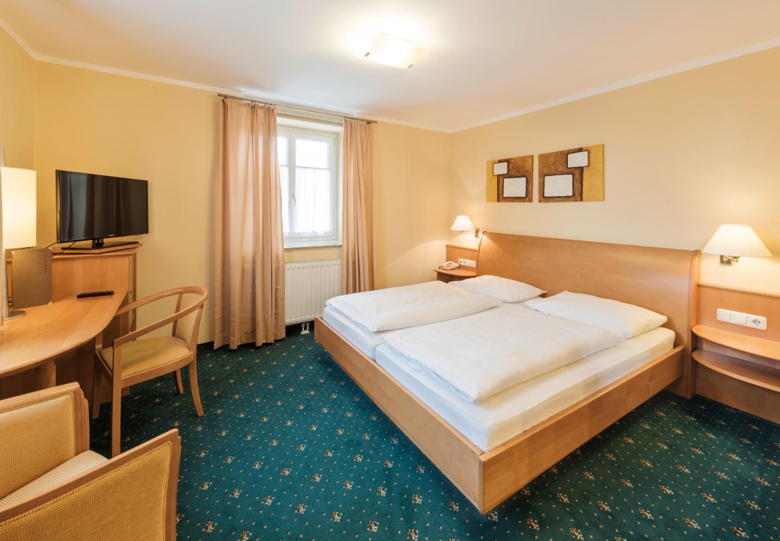A hotel room with a double bed, a desk with a television, a chair, and a window with curtains.