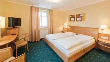 A hotel room with a double bed, a desk with a television, a chair, and a window with curtains.
