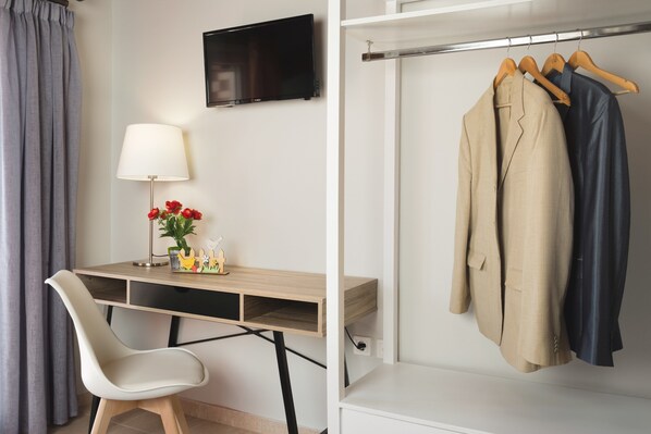A hotel room with a desk, a chair, a TV, and a clothing rack with two jackets.