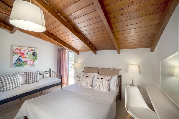 A bedroom with a bed, a bedside table, a lamp, a painting, and a wooden ceiling.