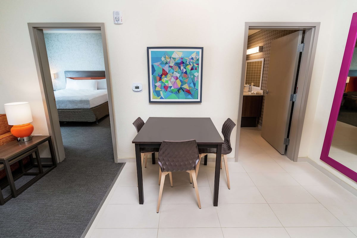 A hotel room with a bed, a dining table with chairs, a painting on the wall, and a bathroom visible through an open door.