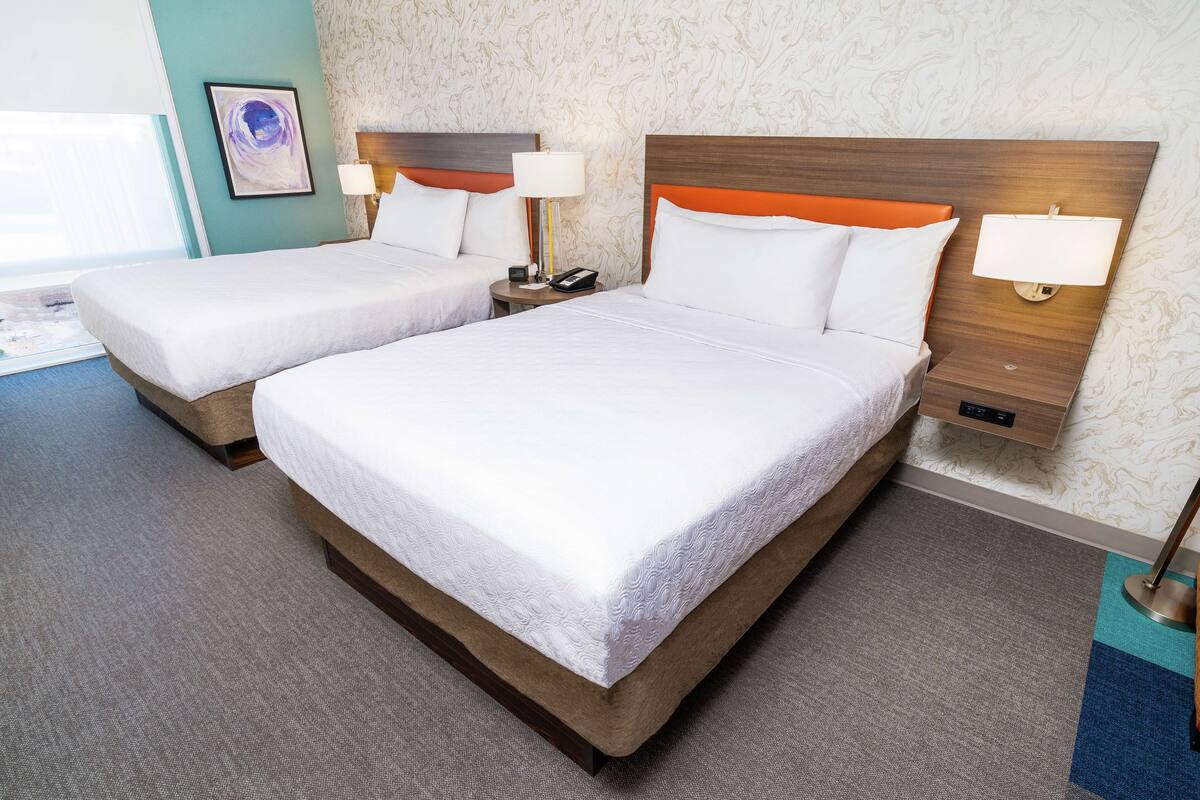 A hotel room with two beds, a bedside table with a lamp, a wall-mounted light, and a patterned wallpaper.