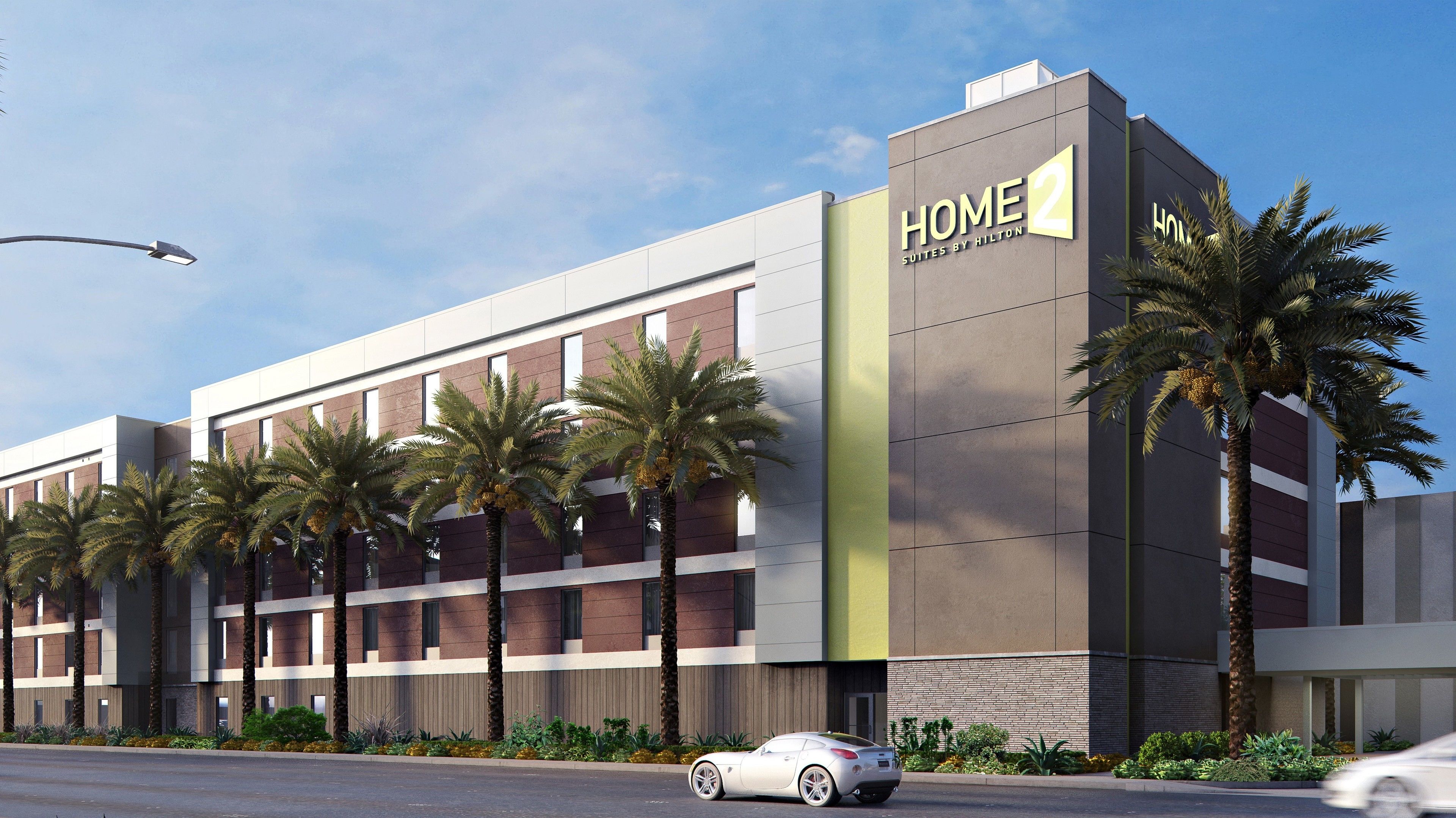 A modern hotel building with a sign reading "HOME 2 Suites by Hilton" and palm trees in front.