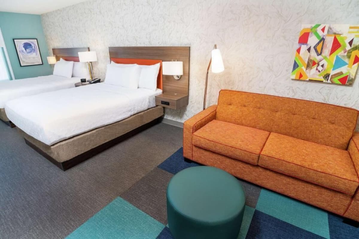 A hotel room with two beds, a sofa, a round ottoman, and a colorful abstract painting on the wall.