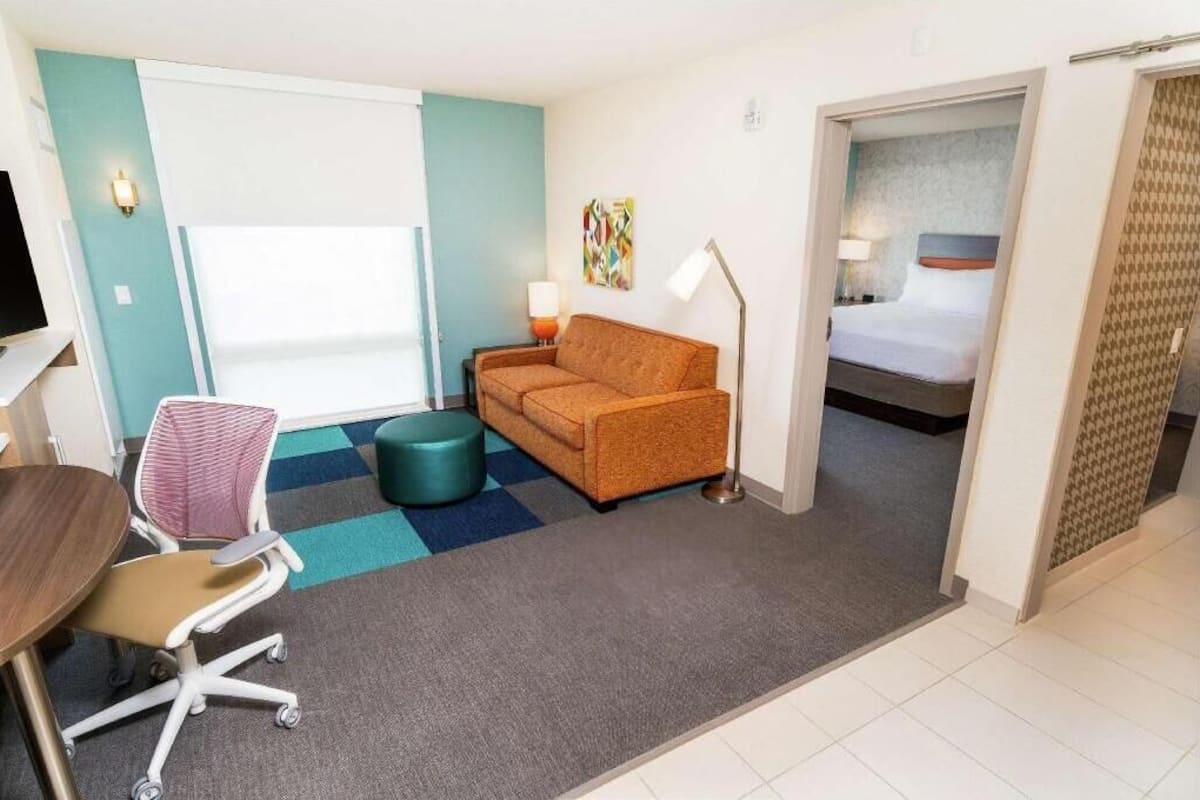A hotel room with a sofa, a desk with a chair, a bed, a lamp, and a colorful abstract painting on the wall.