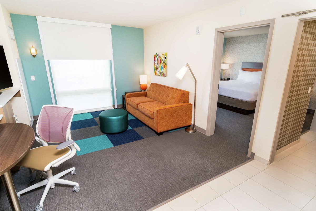 A hotel room with a sofa, a desk with a chair, a small round table, a lamp, and a bed with a headboard.