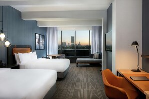 Habitación de hotel con dos camas, un escritorio, una silla, un televisor y vistas al horizonte de la ciudad.