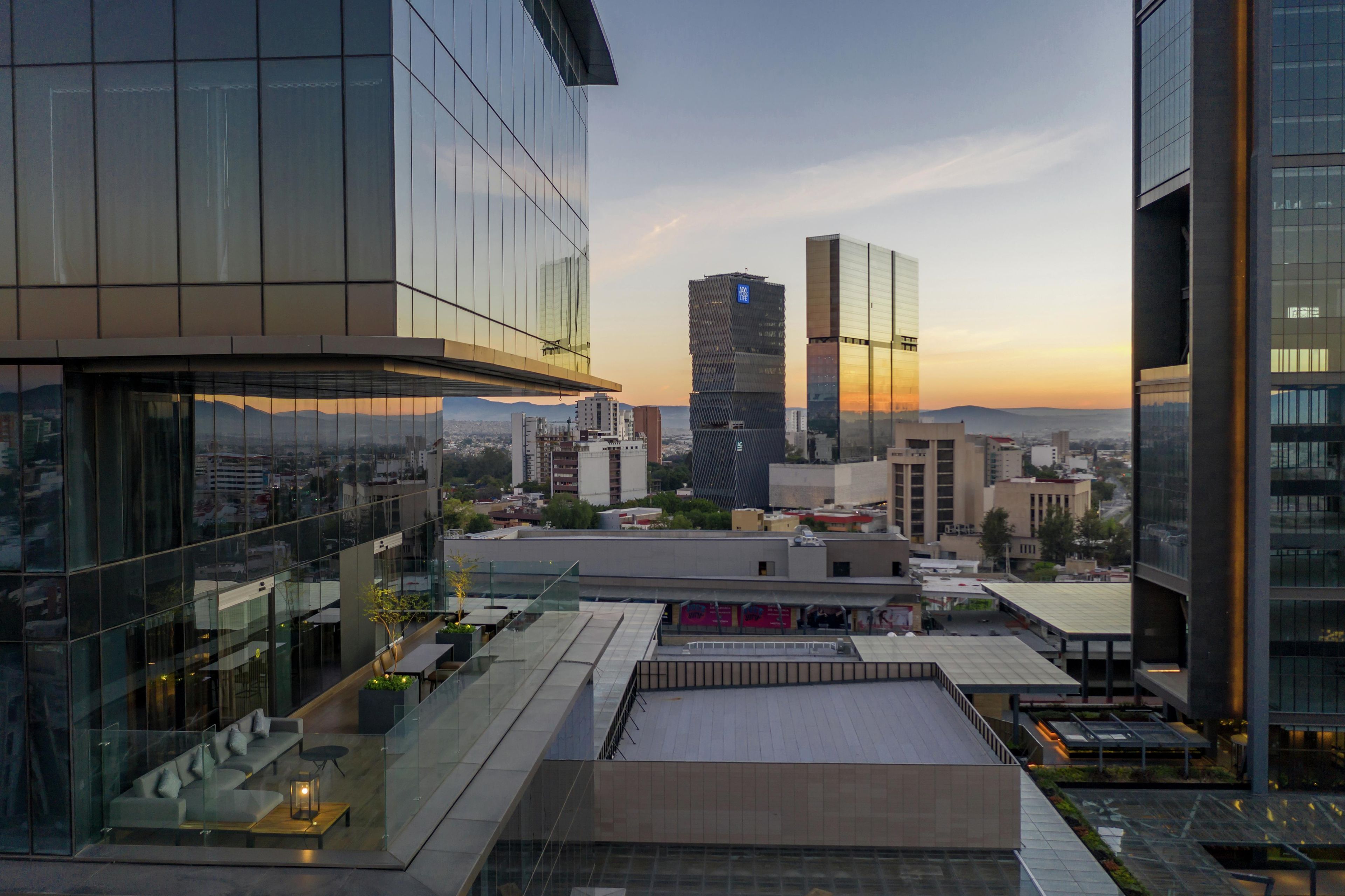 Un edificio moderno con fachada de vidrio, una terraza en la azotea con asientos y vistas a la ciudad, con una torre distintiva al fondo.