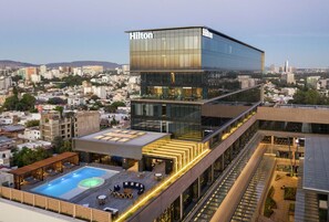 Un moderno hotel Hilton con piscina en la azotea, mobiliario exterior y vistas a la ciudad.