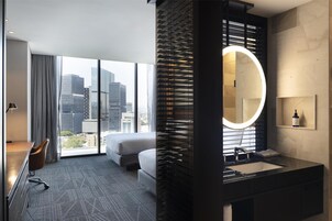 Una habitación de hotel moderna con un ventanal grande, una cama, un escritorio y un baño con un espejo grande y lavamanos.