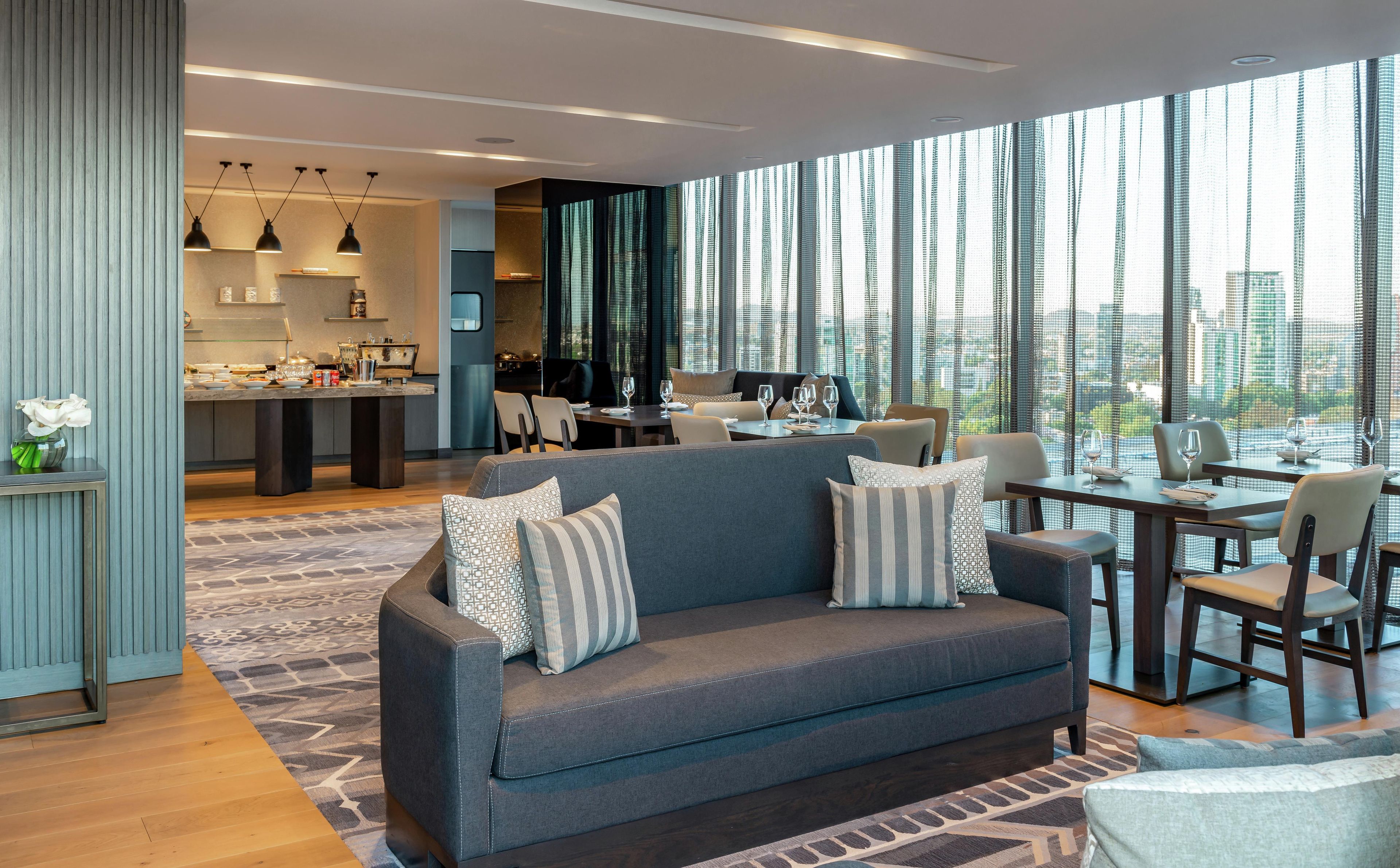 Una sala moderna con un sofá gris, comedor, cocina y ventanas amplias con vista a la ciudad.