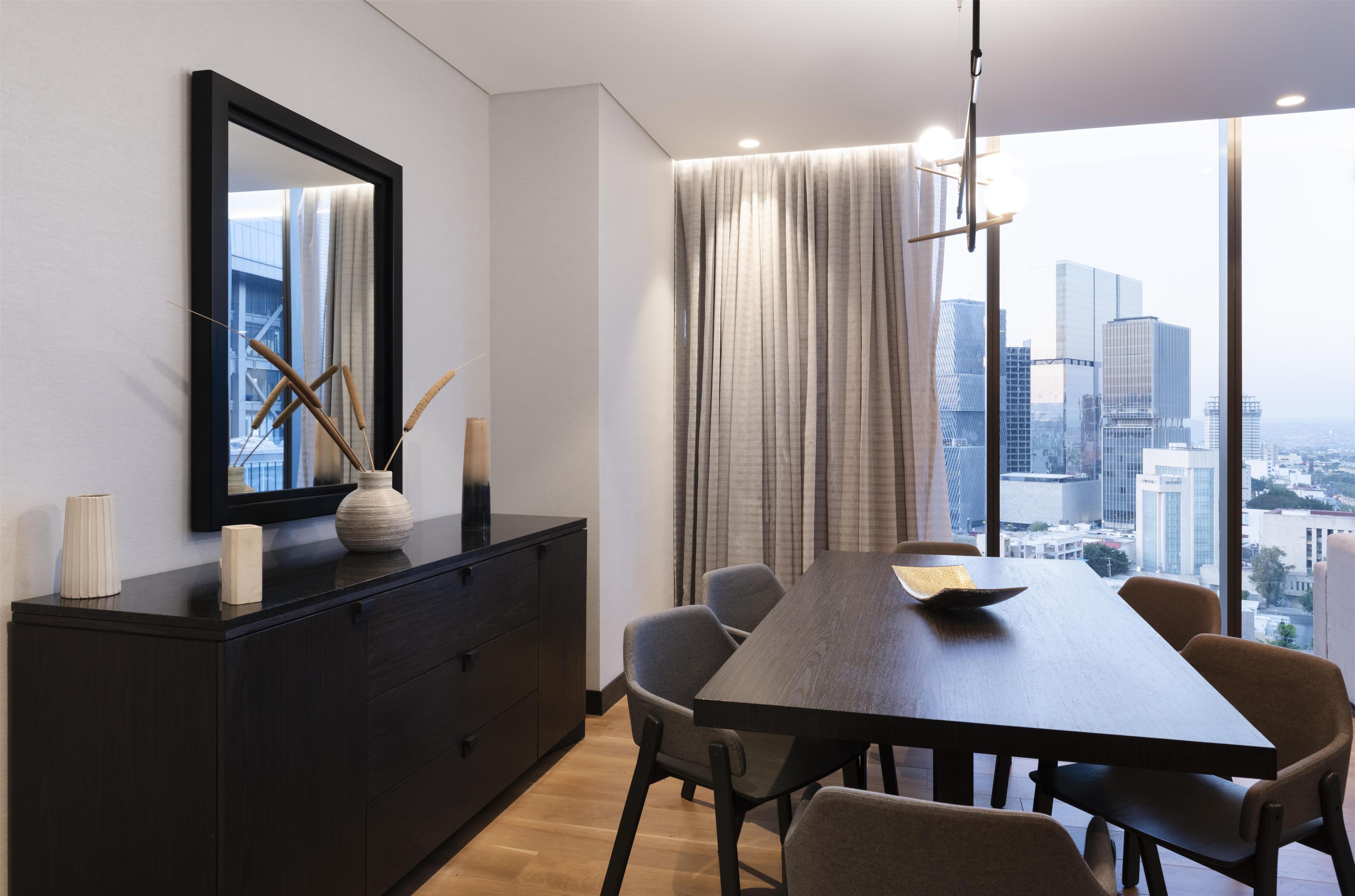 A modern dining area with a dark wooden table, six chairs, a large mirror, and a view of a cityscape through large windows.