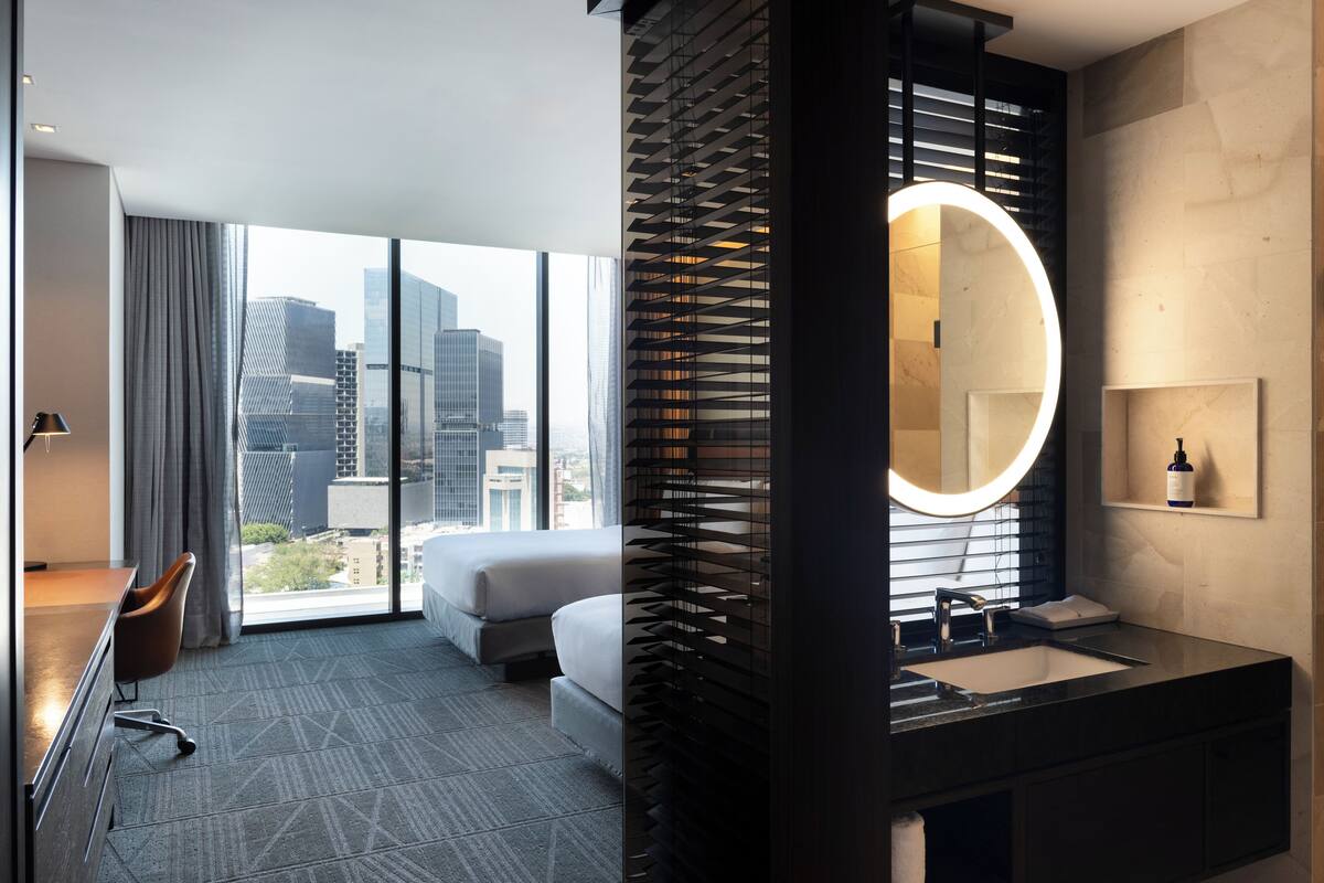 Een moderne hotelkamer met een groot raam, een bed, een bureau en een badkamer met een ronde spiegel en wastafel.