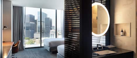A modern hotel room with a large window, a bed, a desk, and a bathroom with a round mirror and sink.