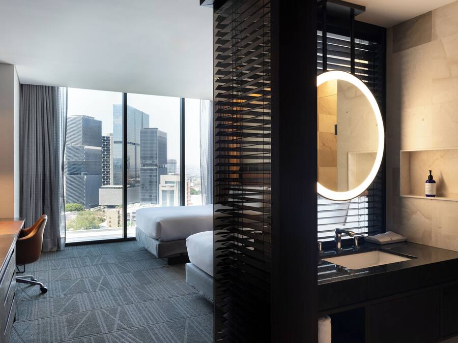 Una habitación de hotel moderna con un ventanal grande, una cama, un escritorio y un baño con un espejo redondo y lavamanos.
