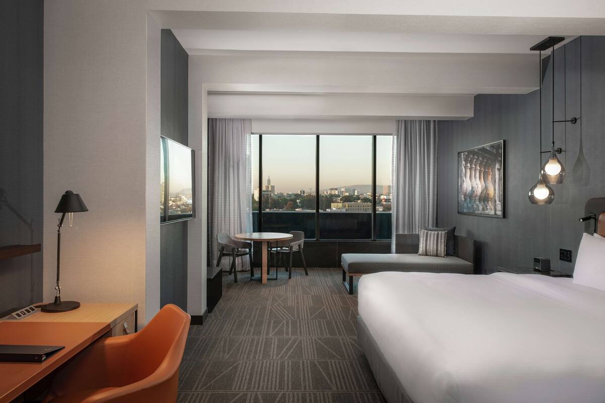 Een hotelkamer met een groot bed, een bureau, een stoel, een kleine tafel en uitzicht op de stad door grote ramen.