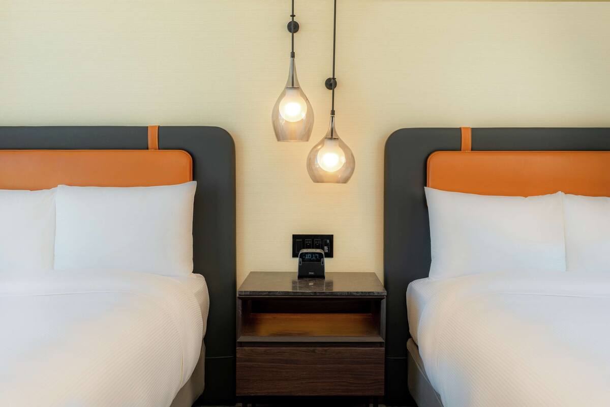 Een hotelkamer met twee bedden, elk met een oranje hoofdbord en witte beddengoed. Twee hanglampen hangen boven de bedden, en een klein houten nachtkastje met een stopcontact en een klok tussen de bedden.