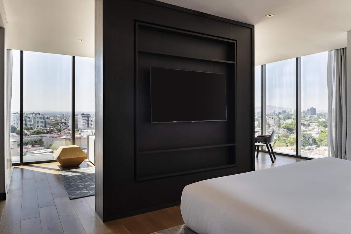 Een moderne slaapkamer met een groot bed, een televisie aan een donkere muur en een stadszicht door grote ramen.