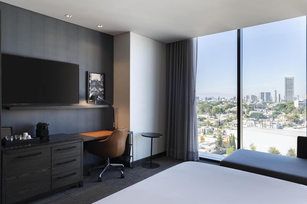 Een moderne hotelkamer met een groot bed, een bureau en een stoel. Grote ramen bieden uitzicht op een stadsbeeld.