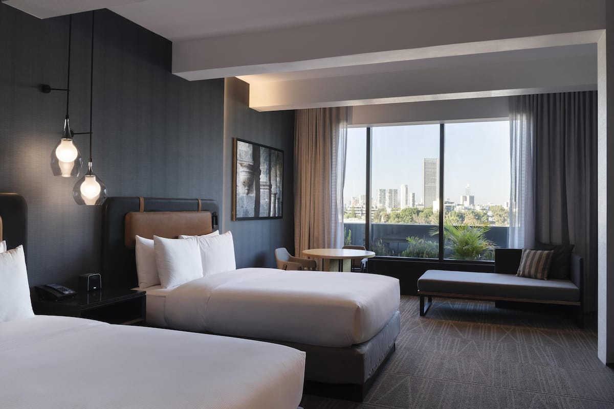 Een hotelkamer met twee bedden, een bank, een kleine tafel en een groot raam met uitzicht op de stad.