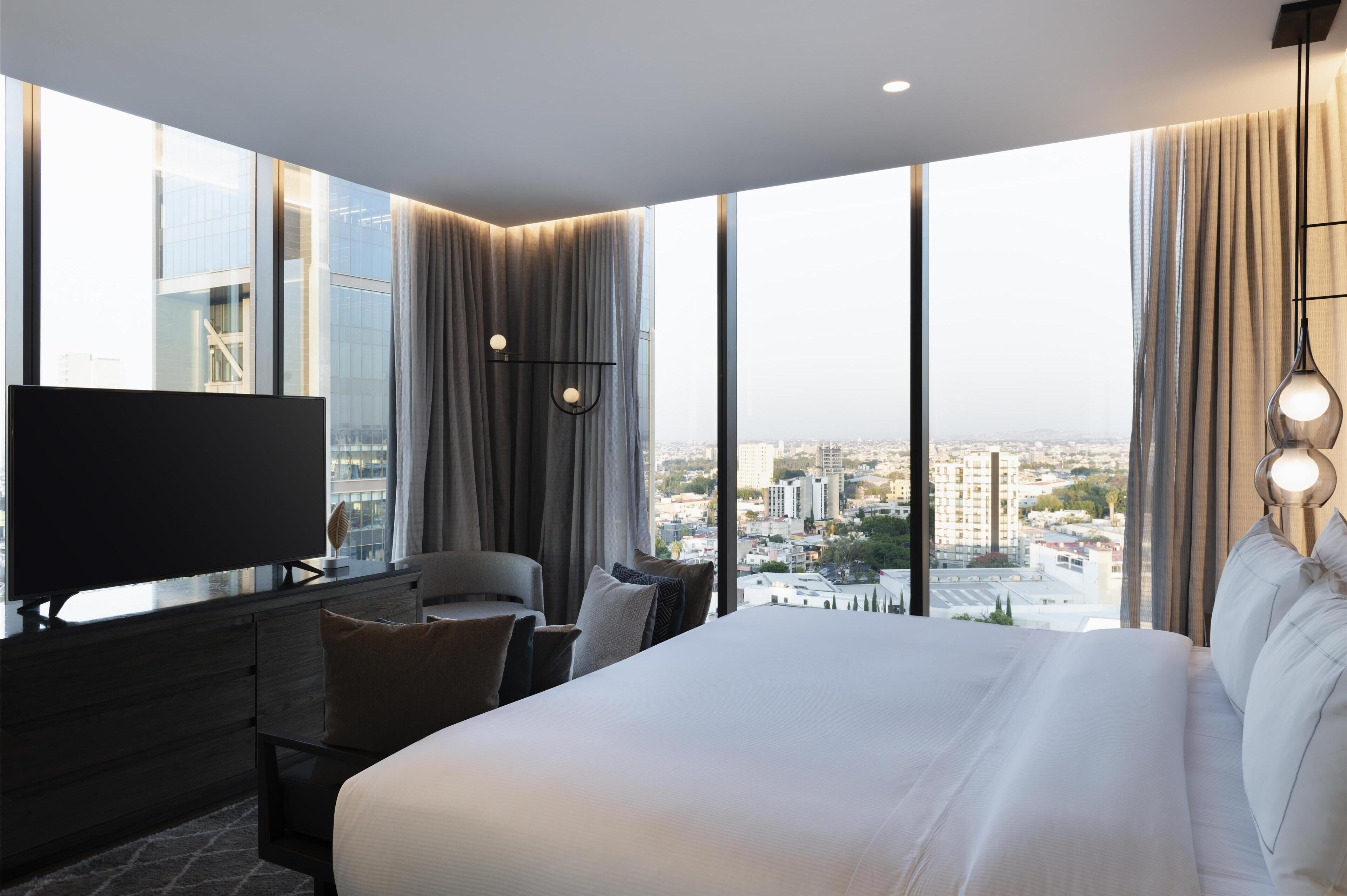 A modern hotel room with a large bed, a desk with a flat-screen TV, and a view of a cityscape through large windows.