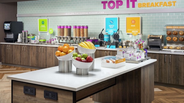 A breakfast buffet area with various food stations, including yogurt, waffles, eggs, and a selection of fruits.