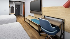 A hotel room with a bed, a desk with a chair, a TV mounted on the wall, and a red lamp.