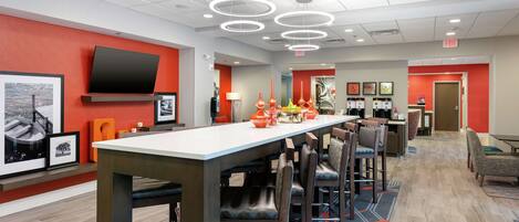 A modern hotel lobby with a long reception desk, multiple chairs, a flat-screen TV, and a variety of decorative items on the walls.