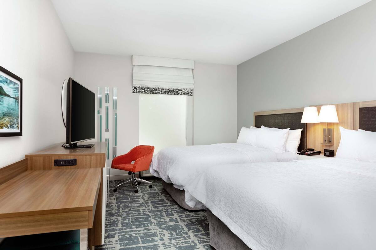 A hotel room with a bed, a desk with a TV, a red chair, a window with blinds, and a nightstand with a lamp.
