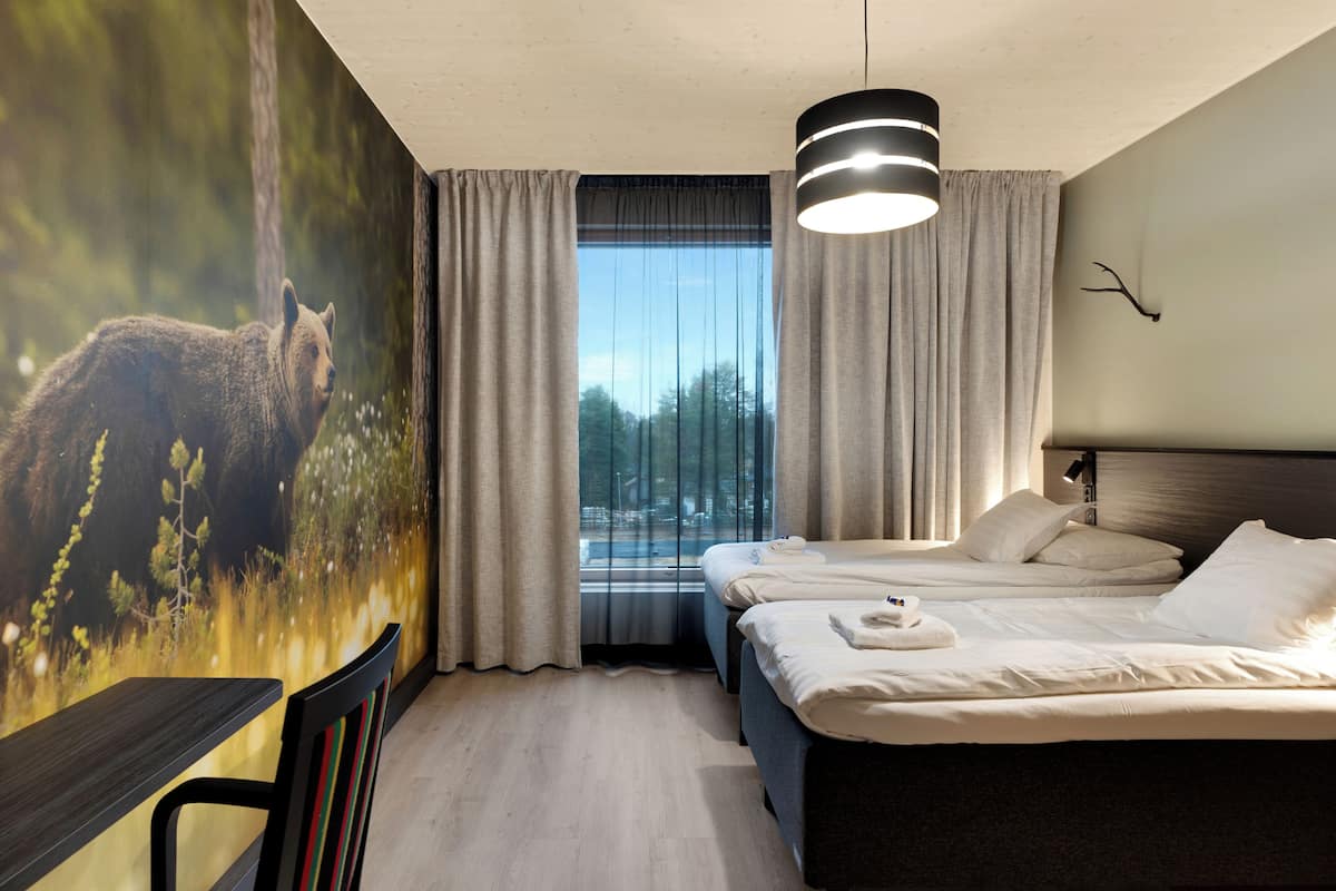 A hotel room with two beds, a large window with curtains, a wall-mounted lamp, and a mural of a bear.