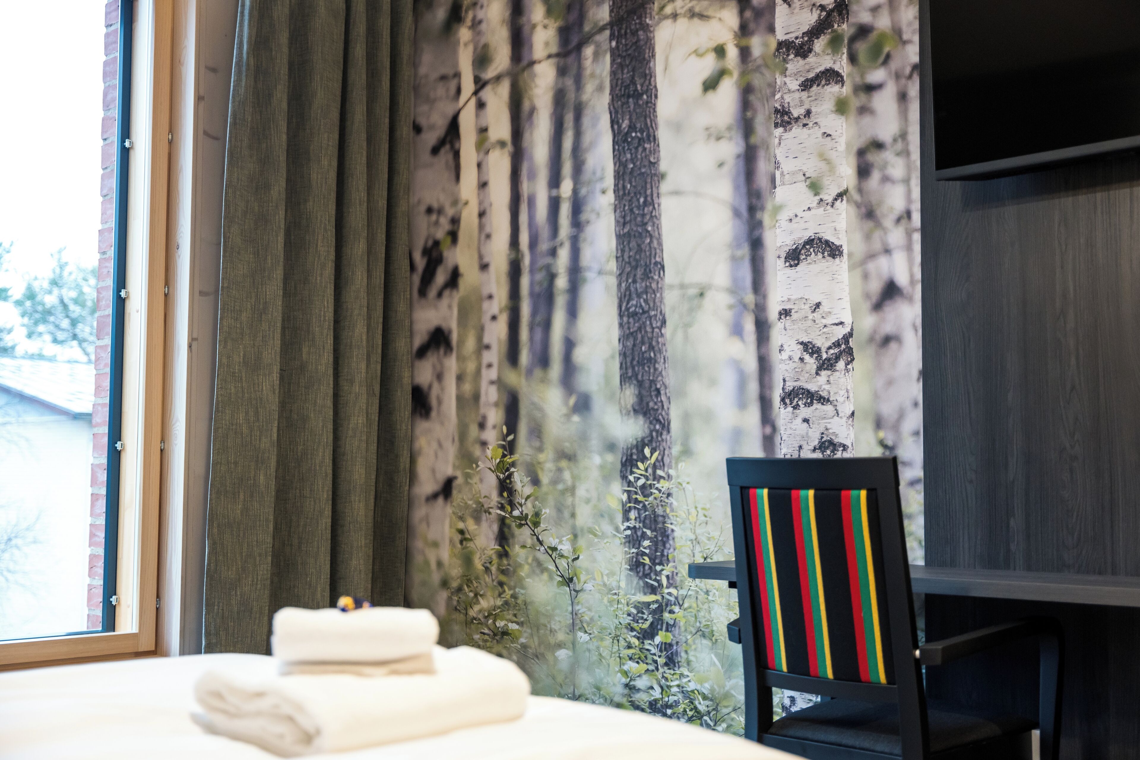 Ett hotellrum med en säng, en stol med en färgglad, randig kudde och utsikt över träd genom ett fönster.