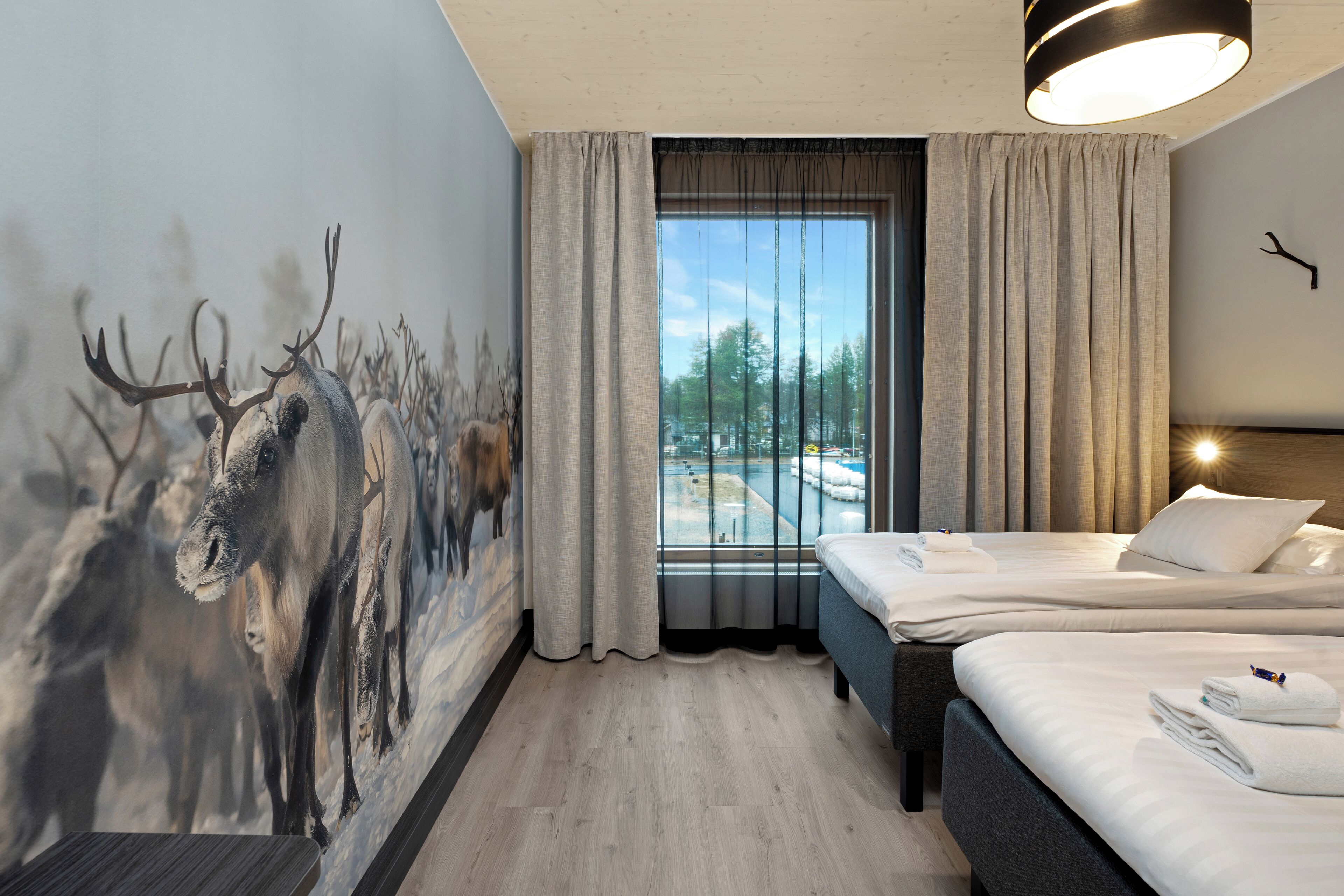 A hotel room with a large window, a bed, a headboard, and a wall mural of deer.
